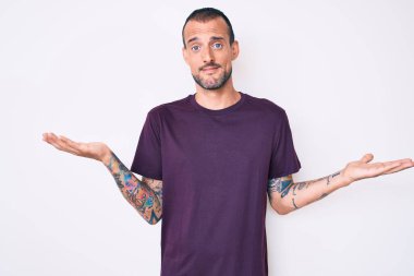 Dövmesi olan, sıradan kıyafetler giyen, kafası karışmış, kolları ve elleri havada genç ve yakışıklı bir adam. şüphe kavramı. 