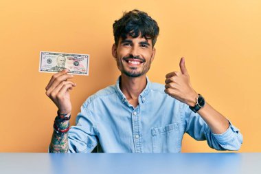 50 dolarlık banknot tutan İspanyol genç adam masaya oturmuş mutlu ve pozitif gülümsüyor, başparmağıyla mükemmel ve onay işareti yapıyor. 
