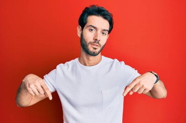 Gündelik beyaz tişört giyen genç İspanyol adam üzgün ve üzgün görünüyor, parmaklarıyla yönü gösteriyor, mutsuz ve depresif.. 