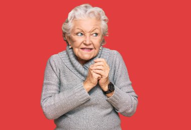 Kış süveteri giyen yaşlı gri saçlı kadın elleri çenesinde gergin ve heyecanlı bir şekilde yan tarafa bakıyor. 