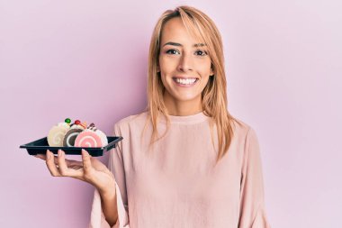 Güzel sarışın kadın elinde pasta tatlısı, pozitif ve mutlu görünüyor. Duruyor ve gülümsüyor. Kendine güvenen bir gülümsemeyle dişlerini gösteriyor. 