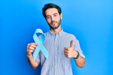 Mavi kurdeleli genç adam mutlu ve pozitif gülümsüyor, başparmağıyla mükemmel ve onay işareti yapıyor. 