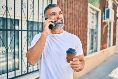 Orta yaşlı, kır saçlı, akıllı telefonla konuşan ve şehirde kahve içen bir adam.