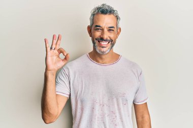 Orta yaşlı, gri saçlı, günlük kıyafetler giyen, gülümseyen, el ve parmaklarıyla 