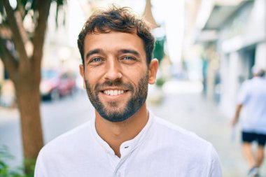 Güneşli bir günde günlük beyaz gömlek giyen sakallı yakışıklı adam dışarıda mutlu bir şekilde gülümsüyor.