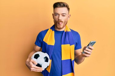 Genç kızıl saçlı, holigan atkısı takan ve akıllı telefon kullanan komik bir ifadeyle dilini dışarı çıkaran adam.. 