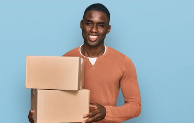 Afro-Amerikalı genç adam teslimat paketini tutuyor. Olumlu ve mutlu görünüyor. Kendine güvenen bir gülümsemeyle dişlerini gösteriyor. 