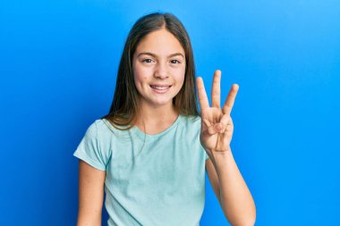 Güzel esmer, beyaz tişört giyen küçük bir kız üç numaralı parmaklarıyla onu gösterirken kendinden emin ve mutlu bir şekilde gülümsüyor.. 