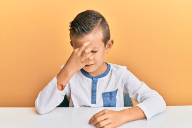 Sevimli beyaz çocuk, gündelik giysiler giyip masada oturuyor. Burnu ve gözleri ovuşturuyor, yorgunluk ve baş ağrısı çekiyor. Stres ve hüsran kavramı. 