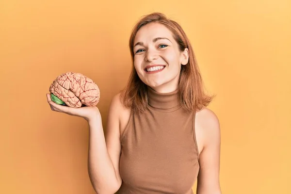Young caucasian woman holding brain looking positive and happy standing and smiling with a confident smile showing teeth