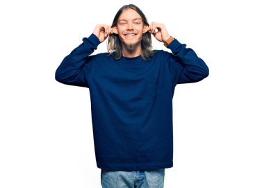 Uzun saçlı, sıradan bir süveter giyen, kulakları parmaklarıyla çeken yakışıklı beyaz bir adam. Komik bir jest. deneme sorunu 