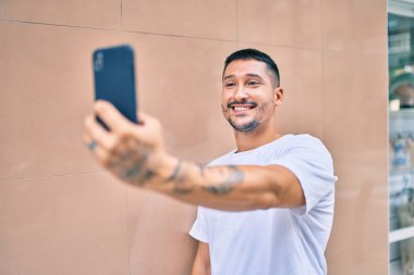 Genç İspanyol gülümseyen adam şehrin caddesindeki akıllı telefonun yanında selfie çekerken mutlu oluyor..