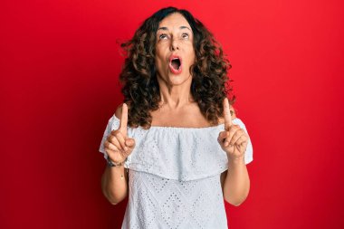 Orta yaşlı İspanyol kadın gündelik kıyafetler giyip şaşkın şaşkın yukarı bakıyor ve parmaklarıyla işaret ediyor ve kollarını kaldırıyor.. 