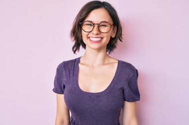 Genç ve güzel bir kadın günlük kıyafetler ve gözlük takıyor. Pozitif ve mutlu görünüyor. Kendine güvenen bir gülümsemeyle dişlerini gösteriyor. 