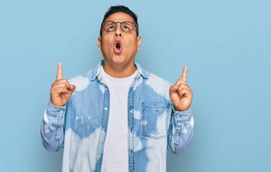 Gündelik kıyafet ve gözlük takan genç latin adam şaşırmış ve şaşırmış bir şekilde yukarı baktı ve parmaklarıyla işaret etti ve kollarını kaldırdı.. 