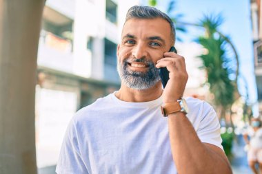 Orta yaşlı, kır saçlı, mutlu gülümseyen bir adam şehirde akıllı telefon kullanıyor.