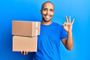 Hispanik yetişkin bir adam mavi arka planda paketler tutuyor tamam işareti yapıyor parmaklarıyla, gülümseyerek mükemmel bir sembol oluşturuyor. 