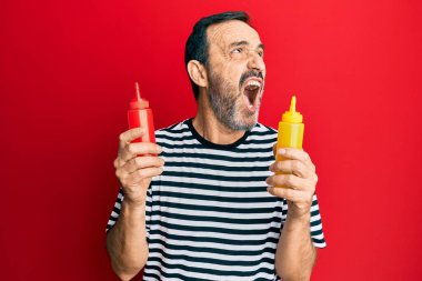 Orta yaşlı Latin bir adam elinde ketçap ve hardal şişesi tutarken sinirli ve öfkeli çığlıklar atıyor. Öfkeyle bağırıyor. öfke ve agresif konsept. 