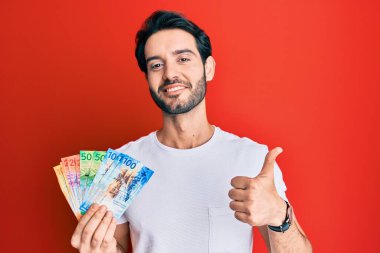 İspanyol asıllı genç adam İsviçre frangı banknotları tutuyor mutlu ve olumlu gülümsüyor, başparmağıyla mükemmel ve onay işareti yapıyor. 