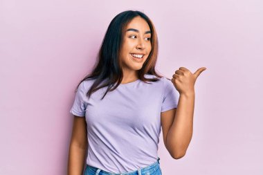 Gündelik giysiler giymiş genç Latin bir kadın başparmağını yana kaldırmış, ağzı açık gülümsüyor. 