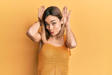 Sarı günlük tişört giyen, tavşan kulağı hareketi yapan, elleri avuçlarında kuşkucu ve alaycı genç, beyaz bir kadın. Paskalya tavşanı konsepti. 