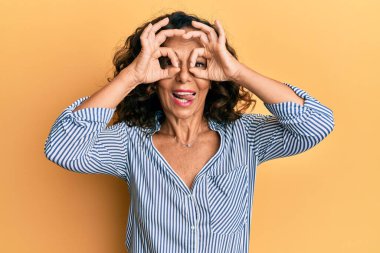 Orta yaşlı İspanyol kadın sıradan kıyafetler giyiyor dürbün gibi jest yapıyor, gözleri parmaklarından bakıyor. çılgın ifade. 