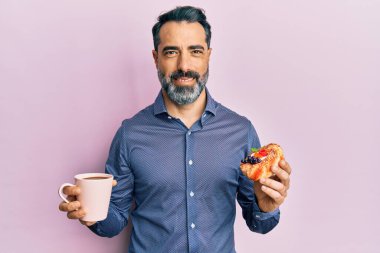 Orta yaşlı, sakallı, kır saçlı kahve içen ve yüzünde mutlu ve havalı bir gülümsemeyle pasta yiyen bir adam. diş gösterme. 