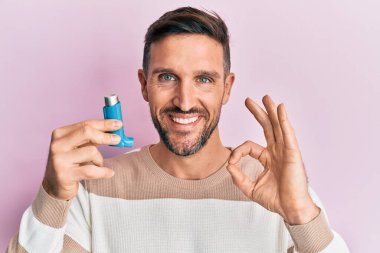 Sakallı yakışıklı adam tıbbi astım spreyi tutuyor. Parmaklarıyla imza atıyor, gülümsüyor. 