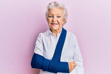 Yaşlı, kır saçlı, kol askısı takan, pozitif ve mutlu görünen, kendine güvenen, gülümseyen, dişlerini gösteren bir kadın. 