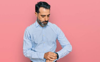 Orta yaşlı, sakallı, iş gömleği giyen, kol saatini kontrol eden, rahat ve kendinden emin bir adam. 