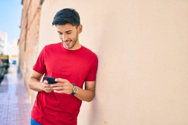 Genç Latin adam şehirde akıllı telefon kullanarak mutlu bir şekilde gülümsüyor..