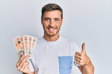 Yakışıklı beyaz adam elinde 10 pound tutuyor mutlu ve olumlu gülümseyen banknotlar, başparmağıyla mükemmel ve onay işareti yapıyor. 