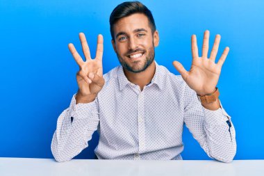 Yakışıklı, iş kıyafetleri giyen, masanın üzerinde oturan ve 8 numaralı parmaklarıyla işaret eden, kendinden emin ve mutlu gülümseyen İspanyol.. 