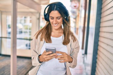 Beyaz kadın şehirde akıllı telefon ve kulaklık kullanarak mutlu bir şekilde gülümsüyor..