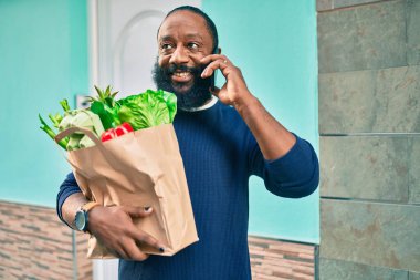 Elinde süpermarketten alınmış kağıt torba sakallı Afrikalı Amerikalı adam telefonda konuşuyor.