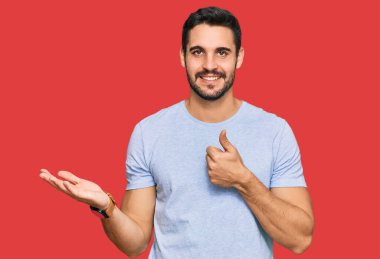 Latin asıllı genç adam, günlük kıyafetler giymiş, avuç içini gösteriyor ve baş parmaklarıyla iyi bir jest yapıyor, mutlu ve neşeli gülümsüyor. 