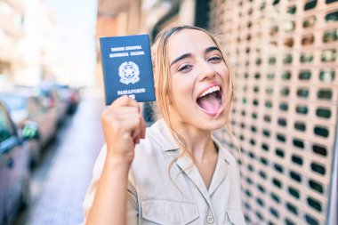 Güneşli bir günde İtalya pasaportunu gösteren güzel sarışın beyaz kadın mutlu bir şekilde dışarıda gülümsüyor.