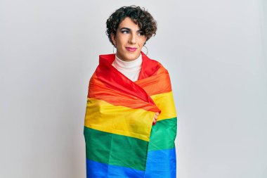 Kadın makyajı yapan genç adam gökkuşağı lgbtq bayrağına sarılmış gülümseyip yan tarafa bakıyor ve düşünüyor.. 