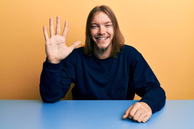 Uzun saçlı, yakışıklı beyaz bir adam masanın üzerine oturmuş, 5 numarayı işaret ediyor ve kendinden emin ve mutlu bir şekilde gülümsüyor.. 