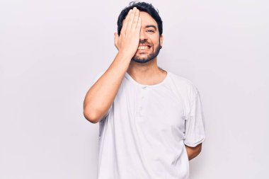 Latin asıllı genç adam gündelik giysiler giyiyor. Bir gözünü eliyle kapatıyor. Yüzünde kendine güvenen bir gülümseme ve sürpriz bir duygu var.. 