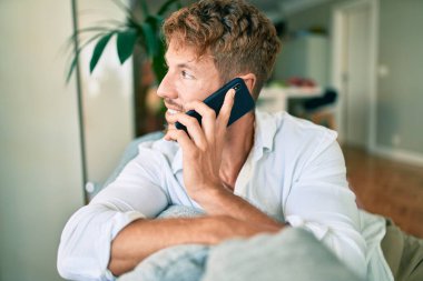 Yakışıklı beyaz adam gülümsüyor. Evdeki kanepede oturup telefonla konuşuyor.