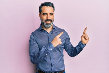 Orta yaşlı, sakallı, gri saçlı iş kıyafetleri giyen, gülümseyen ve kameraya bakan iki eli ve parmakları yanda olan bir adam.. 