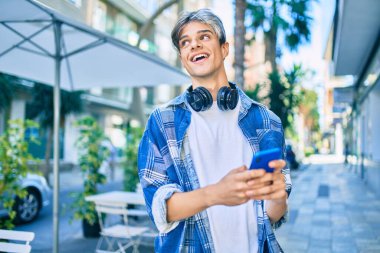 Genç İspanyol adam şehirde akıllı telefon ve kulaklık kullanarak mutlu gülümsüyor..