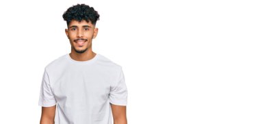 Genç Arap adam, mutlu ve havalı bir gülümsemesi olan günlük beyaz tişört giyiyor. şanslı kişi. 