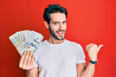 Dolarları tutan İspanyol genç adam başparmağıyla yan tarafa bakıyor mutlu bir şekilde ağzı açık gülümsüyor 