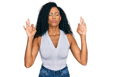 Orta yaşlı Afro-Amerikalı kadın kolsuz gömlekli rahat rahat giyiniyor ve gözleri kapalı gülümsüyor parmaklarıyla meditasyon hareketi yapıyor. yoga konsepti. 