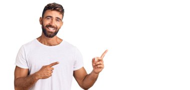 Gündelik beyaz tişört giyen genç İspanyol adam gülümseyip kameraya bakıyor ve iki eliyle ve parmaklarıyla kenarı işaret ediyor.. 