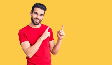 Genç, sakallı, sıradan tişörtlü, gülümseyen ve kameraya bakan iki eli ve parmakları yanda olan yakışıklı bir adam.. 