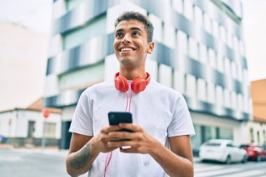 Genç Latin adam şehirde akıllı telefon ve kulaklık kullanarak mutlu bir şekilde gülümsüyor..
