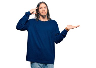 Uzun saçlı yakışıklı beyaz adam, günlük kışlık süveter giyiyor. Kafası karışmış ve avuç içi açık, fotokopi alanı gösteren ve alnını işaret eden biri. Bunu bir düşün.. 